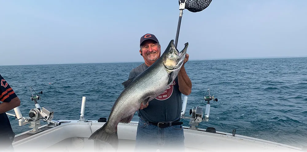 A smiling angler on deck holds up a large king salmon over Lake Michigan